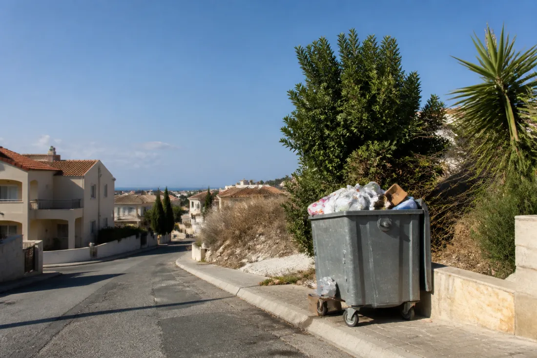 Überfüllter Müllcontainer an einer sonnigen Straße auf Zypern mit Wohnhäusern und mediterraner Landschaft im Hintergrund